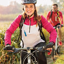 Beautiful happy young woman riding mountain bike over a meadow by the lake or river in the early morning. Young man blurred in the background. Healthy and active sporty lifestyle