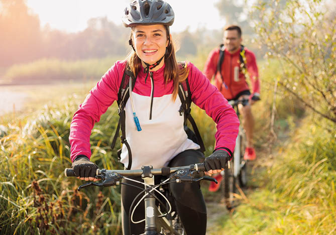 Beautiful happy young woman riding mountain bike over a meadow by the lake or river in the early morning. Young man blurred in the background. Healthy and active sporty lifestyle